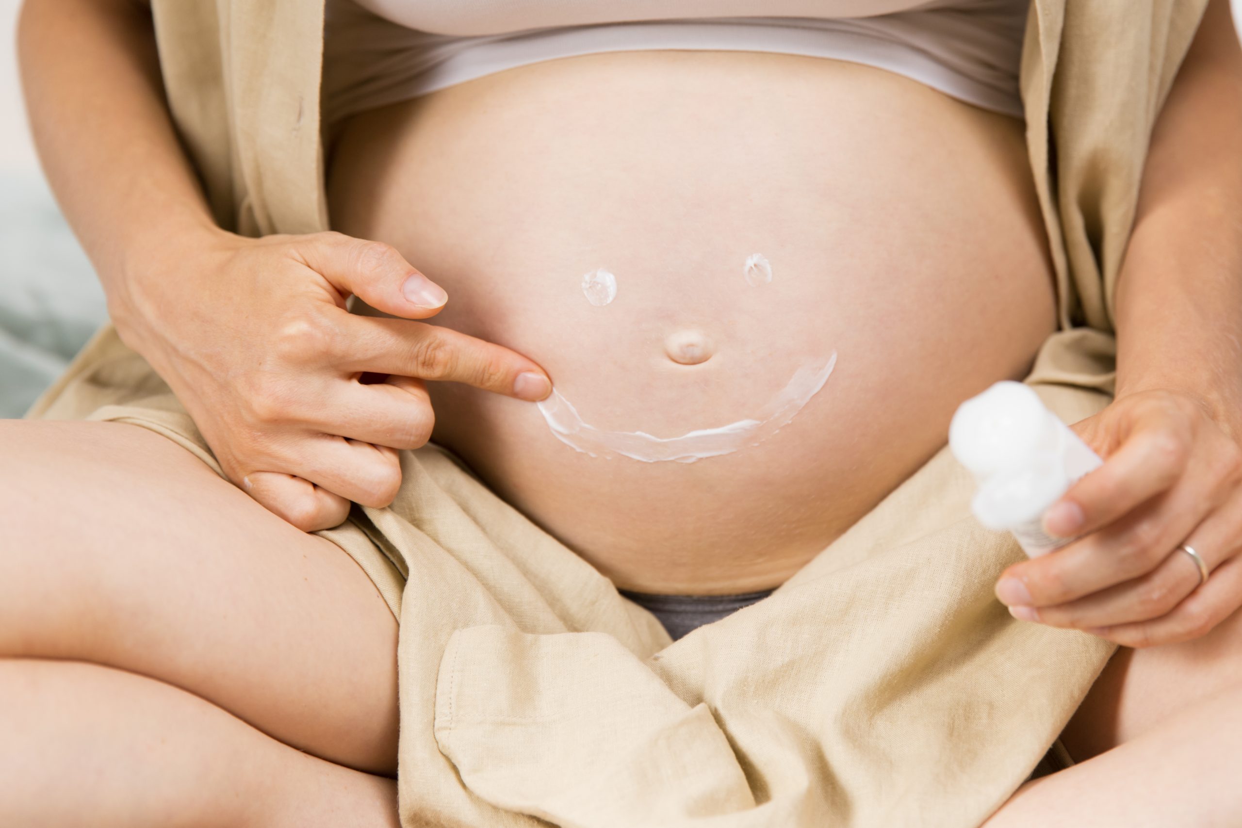 Expectant mother drawing smile with cream on her tummy. Pregnant woman sitting and applying cream to her belly. Pregnancy and body care concept